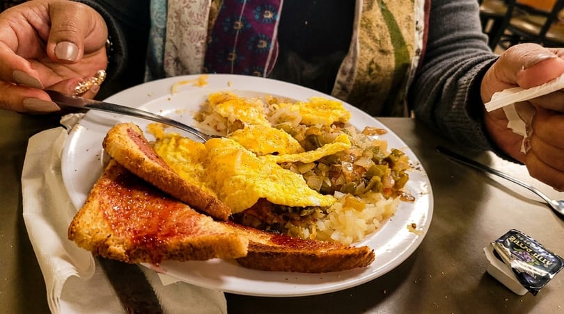 Person eating a breakfast plate of scrambled eggs, hash browns with vegetables, and jam toast.